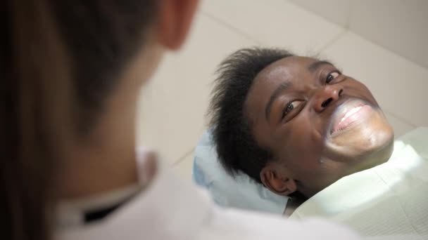 Mooie vrouw arts tandarts. Jonge African American mannelijke patiënt op de leerstoel aan de tandheelkundige kliniek. Geneeskunde, gezondheid, stomatologie concept. tandarts voert inspectie en besluit. op zoek naar de camera — Stockvideo