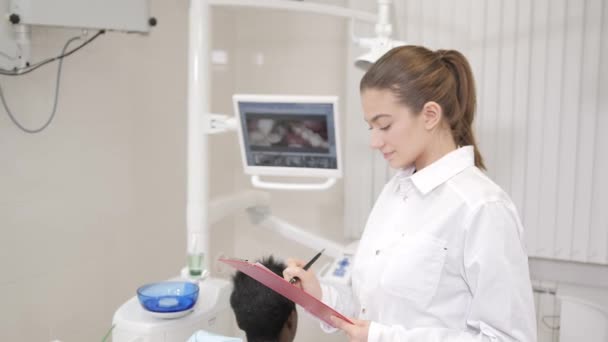 Hermosa doctora dentista. Joven paciente afroamericano en silla en la clínica dental. Medicina, salud, concepto estomatológico. dentista lleva a cabo la inspección y concluye. mirando a la cámara — Vídeo de stock