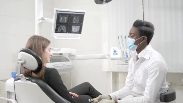 Młody Afroamerykanin lekarz dentysta w maseczkę medyczną. Piękna dziewczyna pacjenta o krzesło w klinice stomatologicznej. Medycyna, zdrowie, Stomatologia koncepcja. dentysta przeprowadza inspekcję i stwierdza — Wideo stockowe