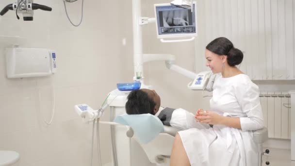 La dentista examina y consulta a la paciente. Joven paciente afroamericano en silla en la clínica dental. Medicina, salud, concepto de estomatología . — Vídeo de stock