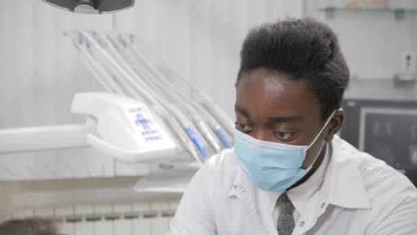 Joven médico afroamericano dentista con máscara médica. Hermosa paciente en la silla de la clínica dental. Medicina, salud, concepto estomatológico. dentista realiza inspección y concluye — Vídeos de Stock