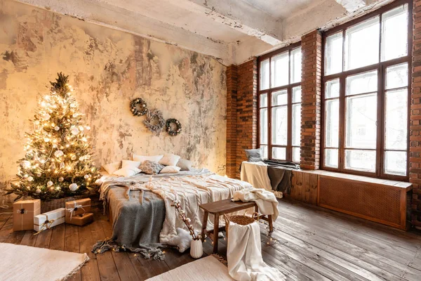 Apartamente în stil Loft. Pat în dormitor, ferestre mari. Zid de cărămidă cu lumânări şi pom de Crăciun. culoare caldă și maro — Fotografie, imagine de stoc