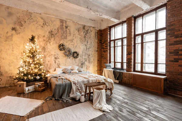 Loft stil lägenheter. Säng i sovrummet, hög stora fönster. Tegelvägg med ljus och julgran. varm och brun färg — Stockfoto