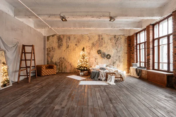 Σοφίτα στυλ διαμερίσματα. Κρεβάτι στην κρεβατοκάμαρα, υψηλή μεγάλα παράθυρα. Τοίχο από τούβλα με κεριά και το χριστουγεννιάτικο δέντρο. ζεστό και καφέ χρώμα — Φωτογραφία Αρχείου