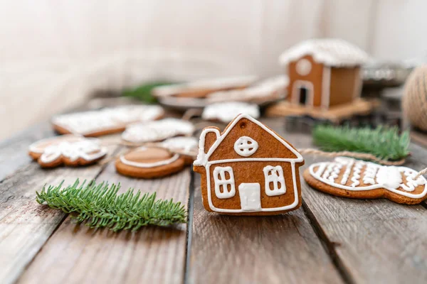 Cute little house in the foreground. A lot of ginger biscuits in different form on brown wooden table. Decorated with white sweet glaze. Christmas mood, winter morning. Fir branches — Stock Photo, Image