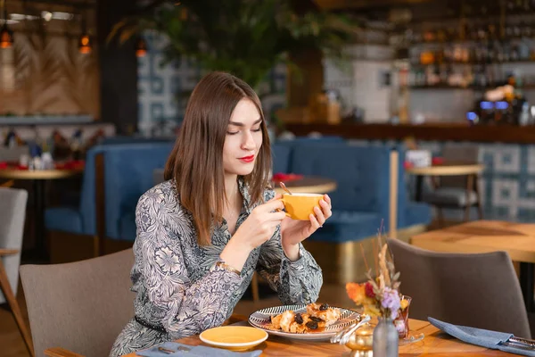 Young woman drinking coffee. Female holding cup. Sitting in coffee shop at wooden table. On table plate with breakfast. pleasant morning, the beginning of the working day — Stock Photo, Image