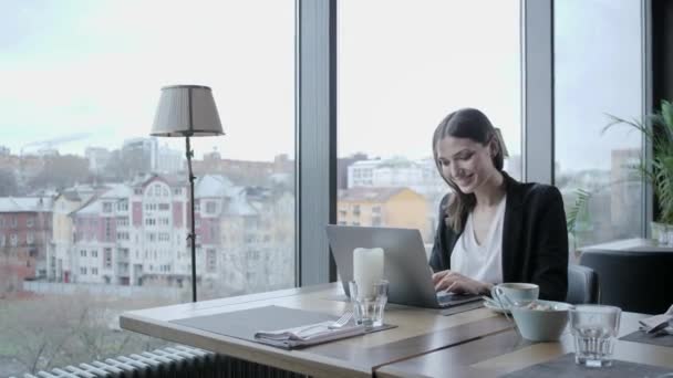 Jonge vrouw lacht, zittend in de coffeeshop op houten tafel. Is grijs aluminium laptop op tafel. Meisje bloggen, surfen op internet, chatten — Stockvideo