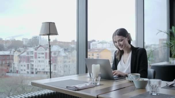 Jovem ri, sentada em uma cafeteria à mesa de madeira. Na mesa está o laptop de alumínio cinza. Girl blogging, navegação na internet, bate-papo — Vídeo de Stock