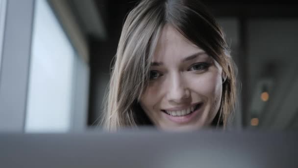 Mujer joven sonrisa y mira en la pantalla del ordenador portátil. Chica blogueando, navegando por Internet, charlando. Se sienta en la cafetería con gran ventana — Vídeos de Stock