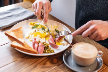 Adam yemek. Bıçak ve çatal elinde. Masaya servis kahvaltı... Amerikan tarzı kahvaltı kızarmış yumurta, sosis, yeşil bezelye ve tost ile. Sağlıklı bir sabah kavramı.