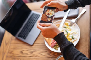 Blogger üstünde smartphone kahvaltı çekiyor. Sosyal ağlar için içerik oluşturma kavramı. Telefonun ekranında odak. Dizüstü bilgisayar ve masada yemek... Amerikan tarzı kahvaltı.