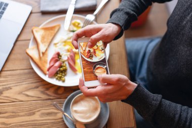 Blogger üstünde smartphone kahvaltı çekiyor. Sosyal ağlar için içerik oluşturma kavramı. Telefonun ekranında odak. Dizüstü bilgisayar ve masada yemek... Amerikan tarzı kahvaltı.