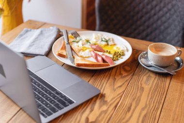 İş yerinde yemek kavramı. Laptop ve masada kahvaltı... Amerikan tarzı kahvaltı kızarmış yumurta, sosis, yeşil bezelye ve tost ile. Sağlıklı bir sabah kavramı.