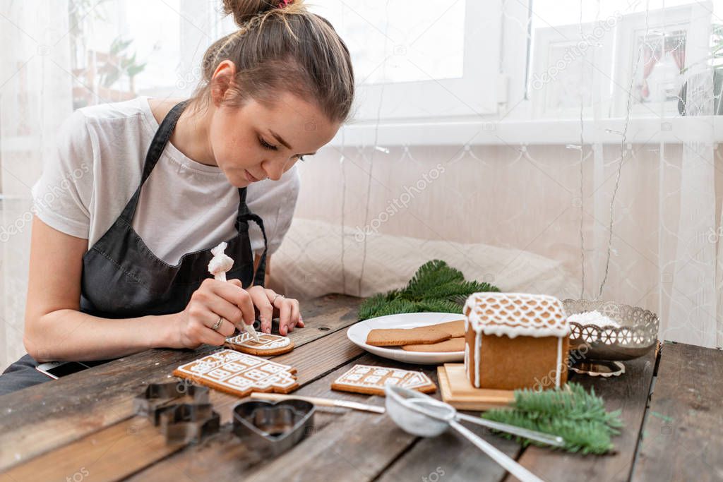 A young girl decorates ginger cookies Christmas winter morning. Woman draws Icing on honey gingerbread cookies. Wooden brown table. copy space. Blank biscuit gingerbread house, ready to decorate.