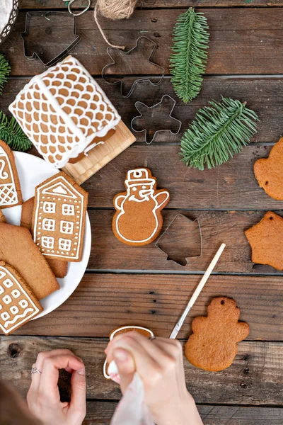 A young girl decorates ginger cookies in the form of snowman Christmas winter morning. Woman draws Icing on honey gingerbread cookies. Wooden brown table. copy space.