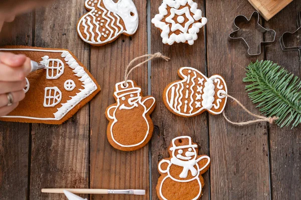 A young girl decorates ginger cookies Christmas winter morning. Woman draws Icing on honey gingerbread house. Wooden brown table. copy space. Blank biscuit gingerbread house, ready to decorate.