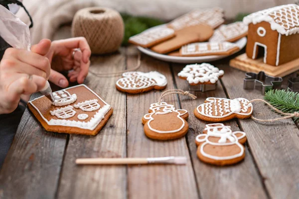A young girl decorates ginger cookies Christmas winter morning. Woman draws Icing on honey gingerbread house. Wooden brown table. copy space. Blank biscuit gingerbread house, ready to decorate.
