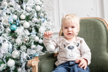 Portre küçük çocuk. Yeni Yılınız Kutlu Olsun. süslü Noel ağacı. Noel sabahı parlak oturma odasında. yeşil sandalye üzerinde oturan