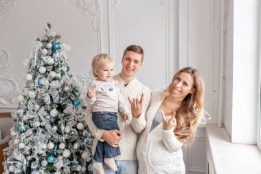 Kameraya dalga geçmeyi. Mutlu aile portresi içinde ana - baba, hamile anne ve küçük oğulları. Yeni Yılınız Kutlu Olsun. süslü Noel ağacı. Noel sabahı parlak oturma odasında.