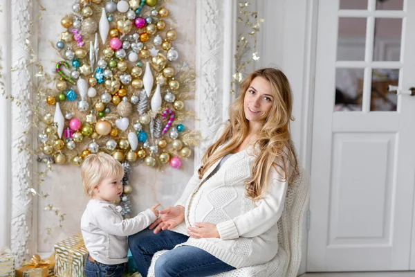 Anne çocukla çalış. Mutlu aile portresi olarak ana sayfa - genç hamile anne küçük oğlunu kucaklar. Yeni Yılınız Kutlu Olsun. süslü Noel ağacı. Noel sabahı parlak oturma odasında. — Stok fotoğraf