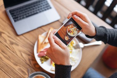 Blogger üstünde smartphone kahvaltı çekiyor. Sosyal ağlar için içerik oluşturma kavramı. Telefonun ekranında odak. Dizüstü bilgisayar ve masada yemek... Amerikan tarzı kahvaltı.