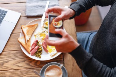 Blogger üstünde smartphone kahvaltı çekiyor. Sosyal ağlar için içerik oluşturma kavramı. Telefonun ekranında odak. Dizüstü bilgisayar ve masada yemek... Amerikan tarzı kahvaltı.