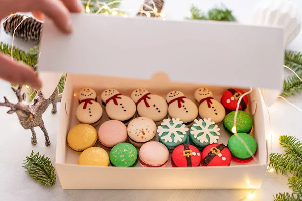 Man opens the lid of the box with his hand. Variety of sweet macaroons in white box. The branches of spruce on bokeh background. Modern european French cuisine. Christmas theme, Merry Christmas card. — Stock Photo, Image