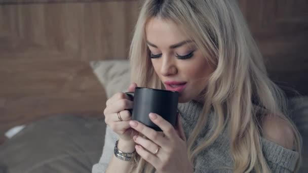 セラミック カップの紅茶やコーヒーを保持している美しい金髪女性をお楽しみください。ベッドの部屋でお茶の時間。見ているウィンドウとお茶を飲む。おはようございます。リラックスできるきれいな若い女の子。幸せな冬のコンセプト. — ストック動画