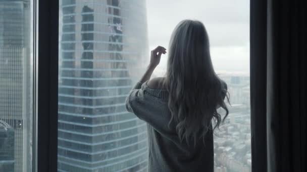 Visszapillantó: gyönyörű szőke nő állt keres teljes hosszúságú ablakon a luxus modern lakás vagy szállodai szoba élvezi téli reggel, és a város a felhőkarcolók Nézd boldog érzés — Stock videók
