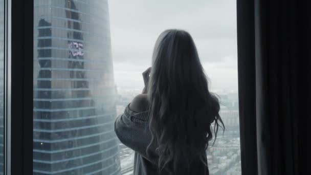 Vista trasera a la hermosa mujer rubia de pie mirando por la ventana de longitud completa de lujo moderno apartamento o habitación de hotel disfrutando de la mañana de invierno y rascacielos de la ciudad ver sentirse feliz — Vídeo de stock