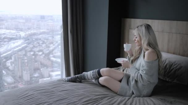 Απολαύστε όμορφη ξανθιά γυναίκα κρατώντας κεραμική κούπα τσάι ή καφέ. Χρόνος τσαγιού στο δωμάτιο κρεβατιών. Κοιτάζοντας από το παράθυρο και να πιείτε καφέ. Καλημέρα. Χαλαρωτική όμορφη νεαρή κοπέλα. Καλό χειμώνα έννοια. — Αρχείο Βίντεο