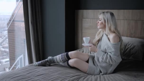 Hermosa mujer rubia sosteniendo taza de cerámica de té o café disfrutar. Hora del té en la cama. Mirando la ventana y bebiendo té. Buenos días. Una chica joven y bonita relajándose. Feliz concepto de invierno . — Vídeo de stock