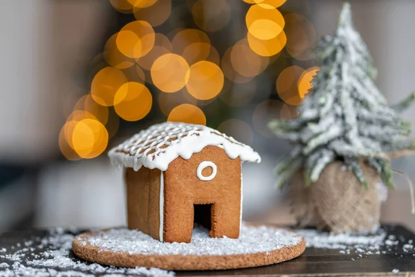 木桌上的姜饼屋。背景上的装饰灯。圣诞树和假日的心情。早晨在明亮的客厅里. — 图库照片