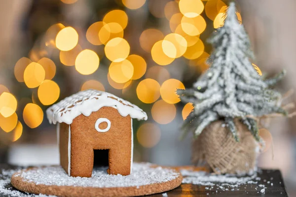 木桌上的姜饼屋。背景上的装饰灯。圣诞树和假日的心情。早晨在明亮的客厅里. — 图库照片