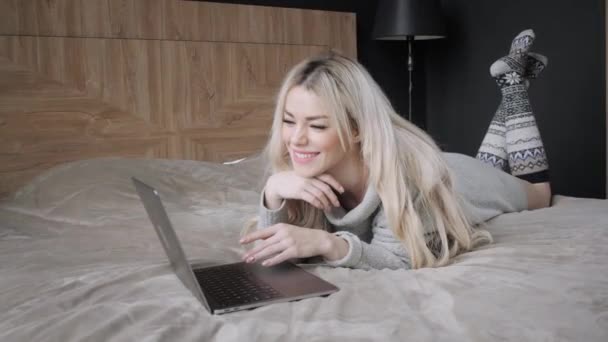 Schöne blonde Frau auf dem Bett liegend mit grauem Aluminium-Laptop. Mädchen lächelt, gute Laune. Bloggen, surfen im Internet, chatten. in einem warmen kuscheligen Pullover und Wollsocken. Frohes Winterkonzept. — Stockvideo