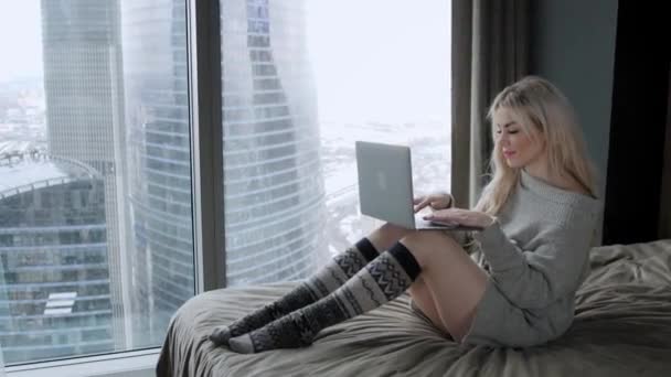 Belle femme blonde assise sur le lit avec un ordinateur portable en aluminium gris. La fille sourit, de bonne humeur. Bloguer, naviguer sur Internet, bavarder. Dans un pull chaleureux et des chaussettes en laine. Concept d'hiver heureux . — Video