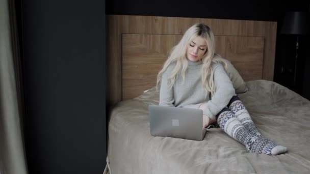 Belle femme blonde assise sur le lit avec un ordinateur portable en aluminium gris. La fille sourit, de bonne humeur. Bloguer, naviguer sur Internet, bavarder. Dans un pull chaleureux et des chaussettes en laine. Concept d'hiver heureux . — Video