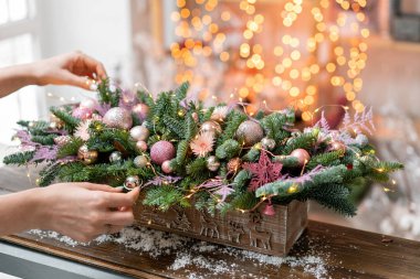 Young woman creates and decorates beautiful festive arrangement of fresh spruce, ornamentals in a rustic wooden box box. Christmas mood. Garland bokeh on background. clipart