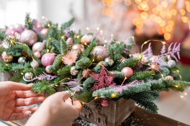 Young woman creates and decorates beautiful festive arrangement of fresh spruce, ornamentals in a rustic wooden box box. Christmas mood. Garland bokeh on background. clipart