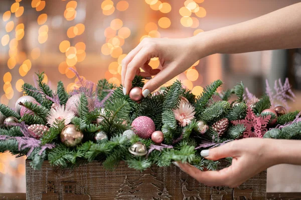 Giovane donna crea e decora bella disposizione festiva di abete rosso fresco, ornamentali in una scatola di legno rustico. L'umore natalizio. Ghirlanda bokeh sullo sfondo . — Foto Stock