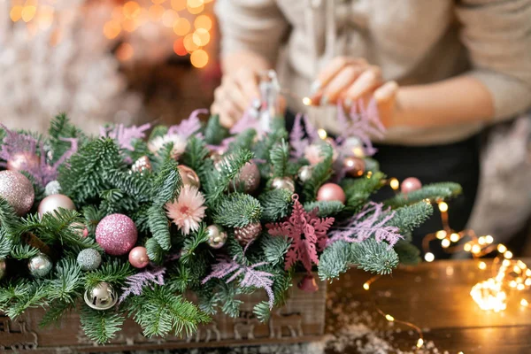 Mujer joven crea y decora hermoso arreglo festivo de abeto fresco, ornamentales en una caja de madera rústica. Humor de Navidad. Guirnalda bokeh en el fondo . — Foto de Stock