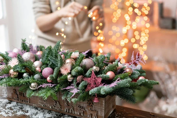 Giovane donna crea e decora bella disposizione festiva di abete rosso fresco, ornamentali in una scatola di legno rustico. L'umore natalizio. Ghirlanda bokeh sullo sfondo . — Foto Stock