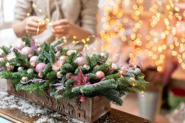 Mujer joven crea y decora hermoso arreglo festivo de abeto fresco, ornamentales en una caja de madera rústica. Humor de Navidad. Guirnalda bokeh en el fondo . — Foto de Stock