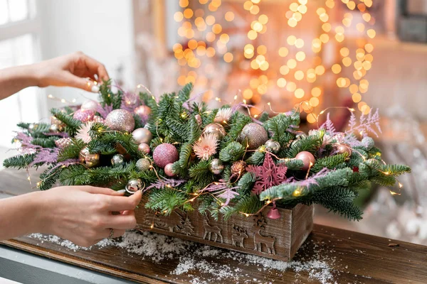 Giovane donna crea e decora bella disposizione festiva di abete rosso fresco, ornamentali in una scatola di legno rustico. L'umore natalizio. Ghirlanda bokeh sullo sfondo . — Foto Stock