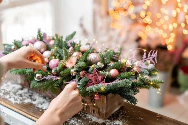 Νεαρή γυναίκα δημιουργεί και διακοσμεί όμορφη εορταστική ρύθμιση από φρέσκο έλατο, καλλωπιστικά φυτά σε ένα ρουστίκ ξύλινο κουτί. Χριστουγεννιάτικη διάθεση. Γιρλάντα bokeh στο παρασκήνιο. — Φωτογραφία Αρχείου
