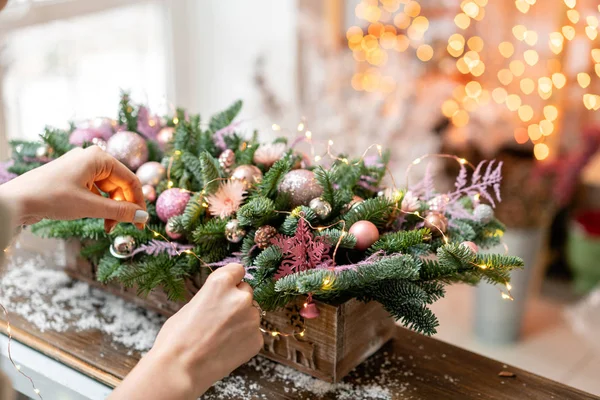 Mulher jovem cria e decora belo arranjo festivo de abeto fresco, ornamentais em uma caixa de caixa de madeira rústica. Humor de Natal. Garland bokeh em segundo plano . — Fotografia de Stock