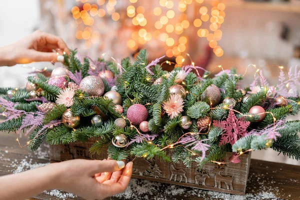 Mujer joven crea y decora hermoso arreglo festivo de abeto fresco, ornamentales en una caja de madera rústica. Humor de Navidad. Guirnalda bokeh en el fondo . — Foto de Stock