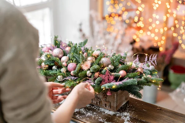 Młoda kobieta tworzy i zdobi piękny układ uroczysty świeżego świerku, rośliny ozdobne w polu rustykalne drewniane pudełko. Świąteczny nastrój. Garland bokeh na tle. — Zdjęcie stockowe