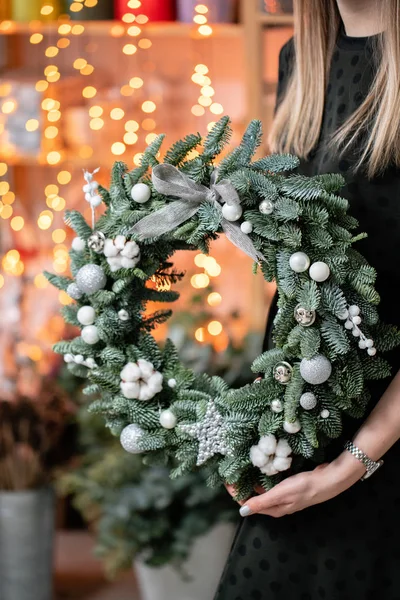 Compras de Navidad. Corona de Navidad verde en manos femeninas. Decorado con elementos grises, algodón y bola de plata. Guirnalda bokeh en el fondo . — Foto de Stock