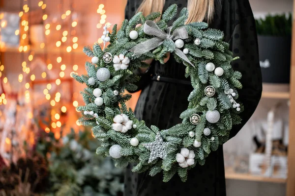 Compras de Navidad. Corona de Navidad verde en manos femeninas. Decorado con elementos grises, algodón y bola de plata. Guirnalda bokeh en el fondo . — Foto de Stock